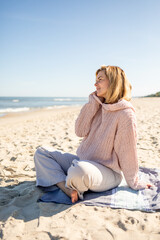 Fototapeta na wymiar Charming young woman in knitted sweater sitting on sandy beach at sunny day