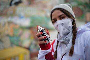 Young female graffiti artist with red nails stands against graffiti wall with her face covered in white hoodie and holds a spray can