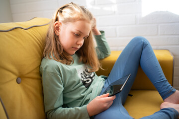cute little girl is sitting on a sofa and enjoying playing an online game on a digital tablet computer, or listening to an online webinar, or reading a book.