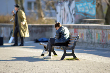 A young woman sits on a park bench while listening to a musician who can be seen blurred in the background. A warm and sunny spring day in Berlin-City.