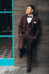 Wedding day. Asian groom poses on the background of a wooden building and large windows.