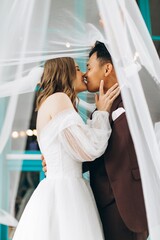 Naklejka premium Wedding day. Beautiful European bride and her Asian groom posing against the backdrop of cafes and large windows.