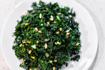 Poster Flat top closeup texture view of green kale vegan vegetarian salad on white plate with pine nuts and olive oil background © Kristina Blokhin