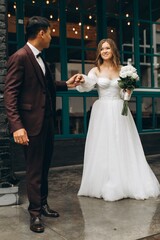 Naklejka premium Wedding day. Beautiful European bride and her Asian groom posing against the backdrop of cafes and large windows.