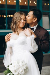 Naklejka premium Wedding day. Beautiful European bride and her Asian groom posing against the backdrop of cafes and large windows.