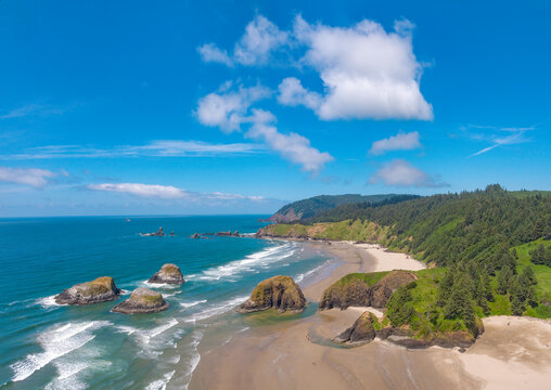 Aerial view on a sunny afternoon Oregon ocean beach with blue water, blue sky, white foam of breaking waves, green grasses and pine trees of Ecola State Park area near Cannon Beach Oregon – color grad