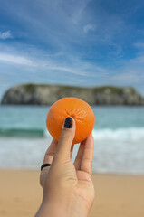 A woman's hand holding a tangerine with the beach in the background