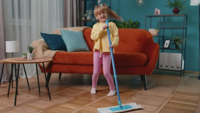 Portrait of carefree Caucasian little adorable child girl washes floors with a mop in domestic living room dancing, singing in positive mood. Happy childhood. Small cute teen school kid at home alone