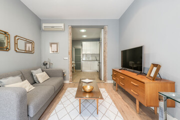 Living room with a three-seater sofa upholstered in gray fabric, a wooden sideboard with drawers and a flat-screen TV, a coffee table and a white kitchen in the background
