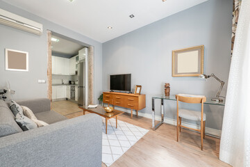 Living room with a three-seater sofa upholstered in gray fabric, a wooden sideboard with drawers and a flat-screen TV, a glass and chromed metal desk and a wooden chair