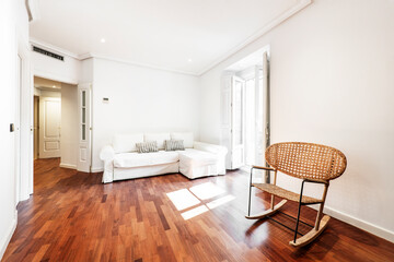 Living room with three-seater white sofa with chaise longue, wicker rocking chair and reddish wood flooring