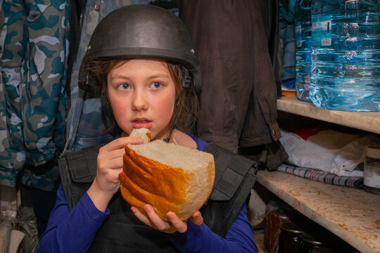 A child in a helmet hides in the basement from the shooting. A girl eats bread in a bomb shelter during the war in Ukraine.