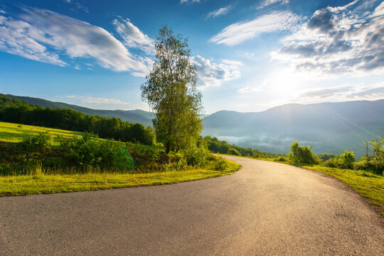 serpentine mountain road down the hill. trees meadow along the way. fog rising in the distant valley. sun above horizon. fluffy clouds on the sky. idyllic travel scenery on a summer morning