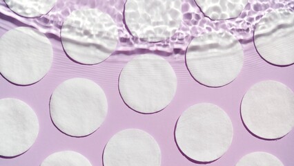 Top view shot of wave coming from above over cotton pads arranged in rows on purple background | Beauty product background