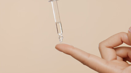 Horizontal close-up shot of clear skin care serum is dripped from dropper on female forefinger on beige background | Skin care serum commercial