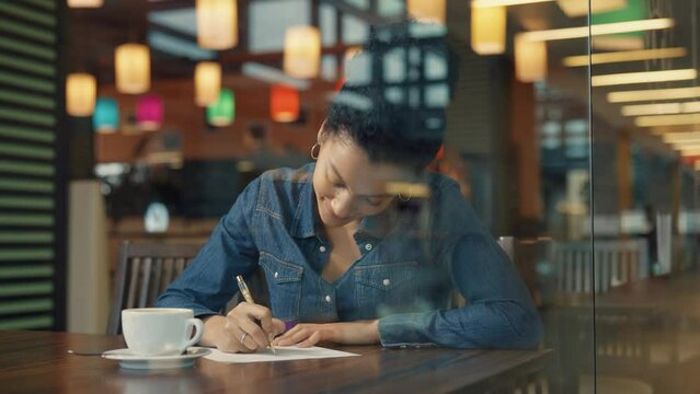 View through a glass window of a young African American woman dreamily thinking about something and then squeaking on a piece of paper. Woman enjoying coffee in a mall cafe. Close. Slow motion.