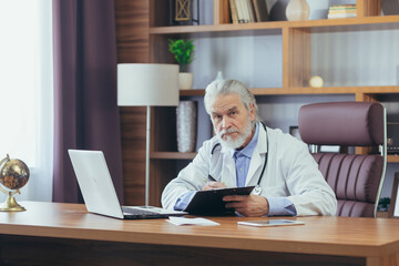 Gray-haired experienced male doctor working on laptop remotely advising patients while sitting in office