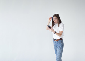 A young cute girl with glasses looks at her mobile phone with a surprised face, standing on a white background