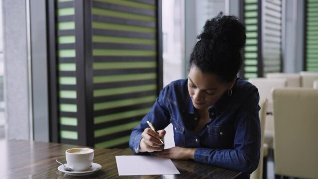 Young African American woman dreamily thinking about something and then squeaking on a piece of paper. Woman enjoying coffee in a mall cafe. Shopping, planning, dreams. Close. Slow motion.