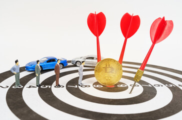 On the target are miniature figures of people, bitcoin, darts. Cars are in the background.