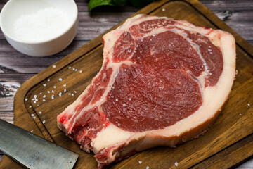 A bone-in steak or t-bone or porterhouse raw on a cutting board on a wooden table with salt and knife. Top view. Close-up.