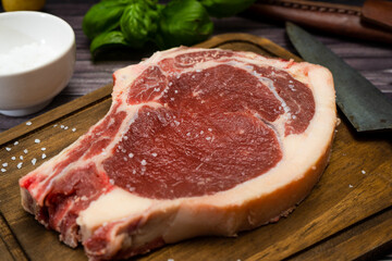 A bone-in steak or t-bone or porterhouse raw on a cutting board on a wooden table with one knfe and salt. High view. Close-up.