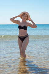 Fototapeta premium A mature slender woman in a swimsuit and a hat is standing in the sea water, smiling. Sea vacation and travel.