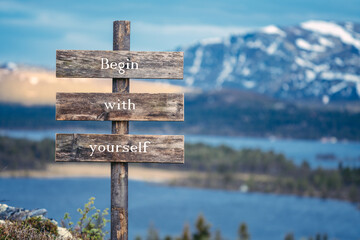 begin with yourself text quote written on wooden signpost outdoors in nature with lake and mountain scenery in the background. Moody feeling.