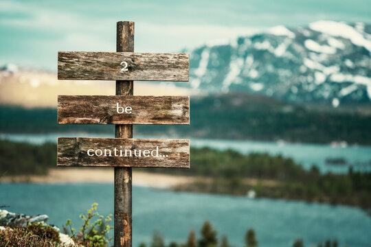 2 be continued text quote written on wooden signpost outdoors in nature with lake and mountain scenery in the background. Moody feeling.
