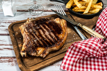 Incredible juicy t-bone cooked on the grill or barbecue on a wooden board accompanied by a french fries and with cutlery on the side.