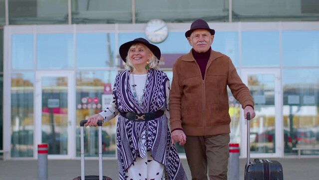 Senior couple happy pensioner tourists grandmother grandfather walking while carrying luggage suitcases on wheels from international airport hall or railway station. Travel, vacation, journey, trip