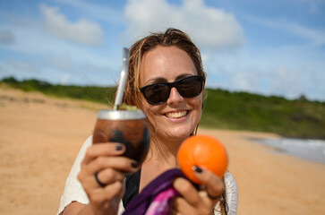 Young woman smiling, holding a yerba mate gourd and a tangerine on a sunny beach day