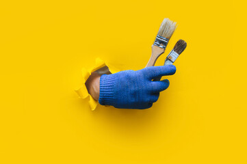 The painter's hand in a blue knitted cloth glove holds two bristle brushes and shows a big stick up...