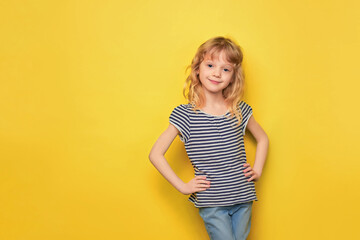 Close-up portrait of a cute attractive cheerful little girl of preschool age. Joyful and smiling child in a striped t-shirt and jeans. Blonde 5 years old on a yellow background with space for text.
