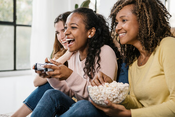 United multiracial young women playing video games wit console at home - Diverse female friends...