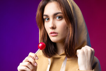 Portrait of a woman in a hoodie, girls licking a red round lollipop with beautiful makeup on a...