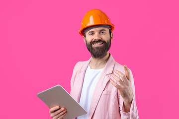 Portrait of young construction engineer wear orange hard hat, in a pink jacket standing on red studio background. A man with a tablet in his hands.
