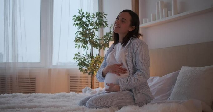 Happy woman caressing her pregnant belly sitting on bed at home