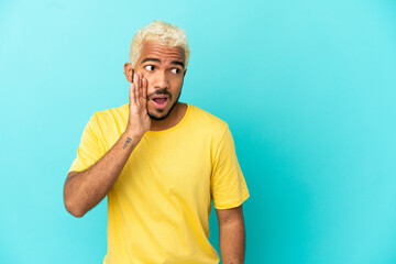 Young Colombian handsome man isolated on blue background whispering something with surprise gesture...