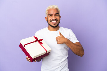 Young Colombian man holding a gift isolated on purple background giving a thumbs up gesture