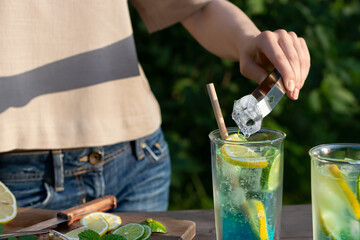Girl makes summer refreshing cocktails blue lagoon. Close-up of adding ice cubes to glasses....