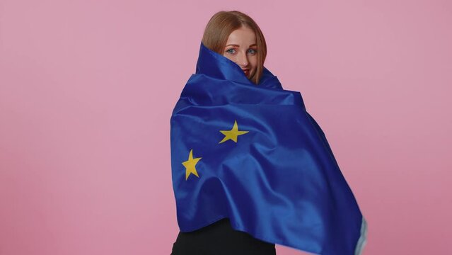 Pretty teen girl in beige blouse waving European Union flag, smiling, cheering democratic laws, human rights freedoms in Europe. Young adult woman indoor isolated on pink studio wall background
