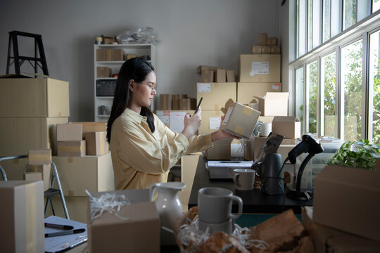 Asian Female Small Business Owner Using Mobile App On Smartphone Checking Parcel Box. Warehouse Worker, Seller Holding Phone Scanning Retail Dropshipping Package Postal Parcel Bar Code.