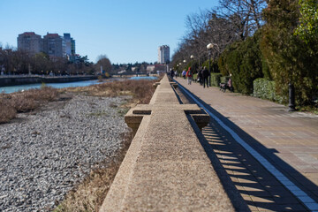 Concrete railings on the embankment of the river. A beautiful shadow falls on the path next to the river. People are walking along the sidewalk. blurry objects. Selective focus..