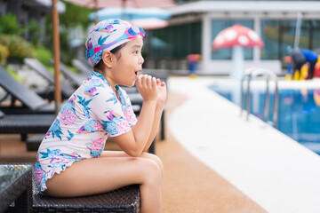 Asian child holiday or kid girl wearing swimsuit to sitting on pool bed and happy smile excited on swimming pool or waterpark for learning swim to exercise on summer school or vacation travel at hotel