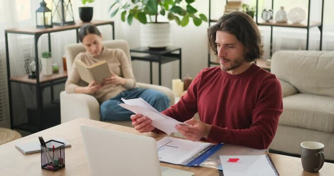 Man working on desk with wife reading book on armchair at home