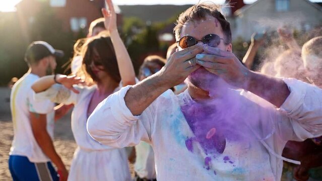 Handsome man claps hands with colored powder in and paints his moustache with lavender-blue dye at Holi festival. People celebrate hindu holiday. End of lockdown, covid pandemic, restrictions.