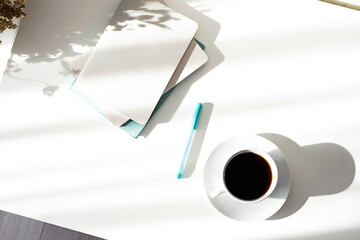 Black coffee cup, stack of notebooks or planners on white table with interesting shadows. Scandinavian style, stationary still life in the morning rays of sun. 