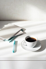 Black coffee cup, stack of notebooks or planners on white table with interesting shadows. Scandinavian style, stationary still life in the morning rays of sun. 