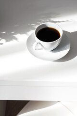 Aromatic black coffee cup on white table with interesting shadows.Scandinavian style still life in the morning rays of sun.